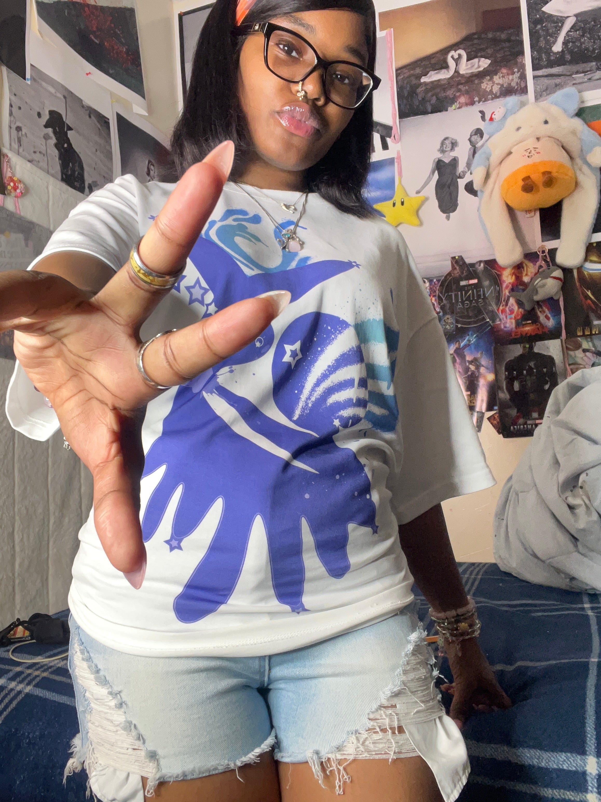 A person wearing a white graphic t-shirt with a blue space-themed print and light-wash distressed denim shorts, posing in a bedroom setting.