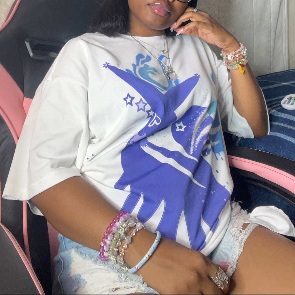 A woman wearing a white graphic t-shirt with a blue and purple design, paired with white shorts, sitting in a pink and black gaming chair against a wall decorated with posters and a yellow pom-pom.