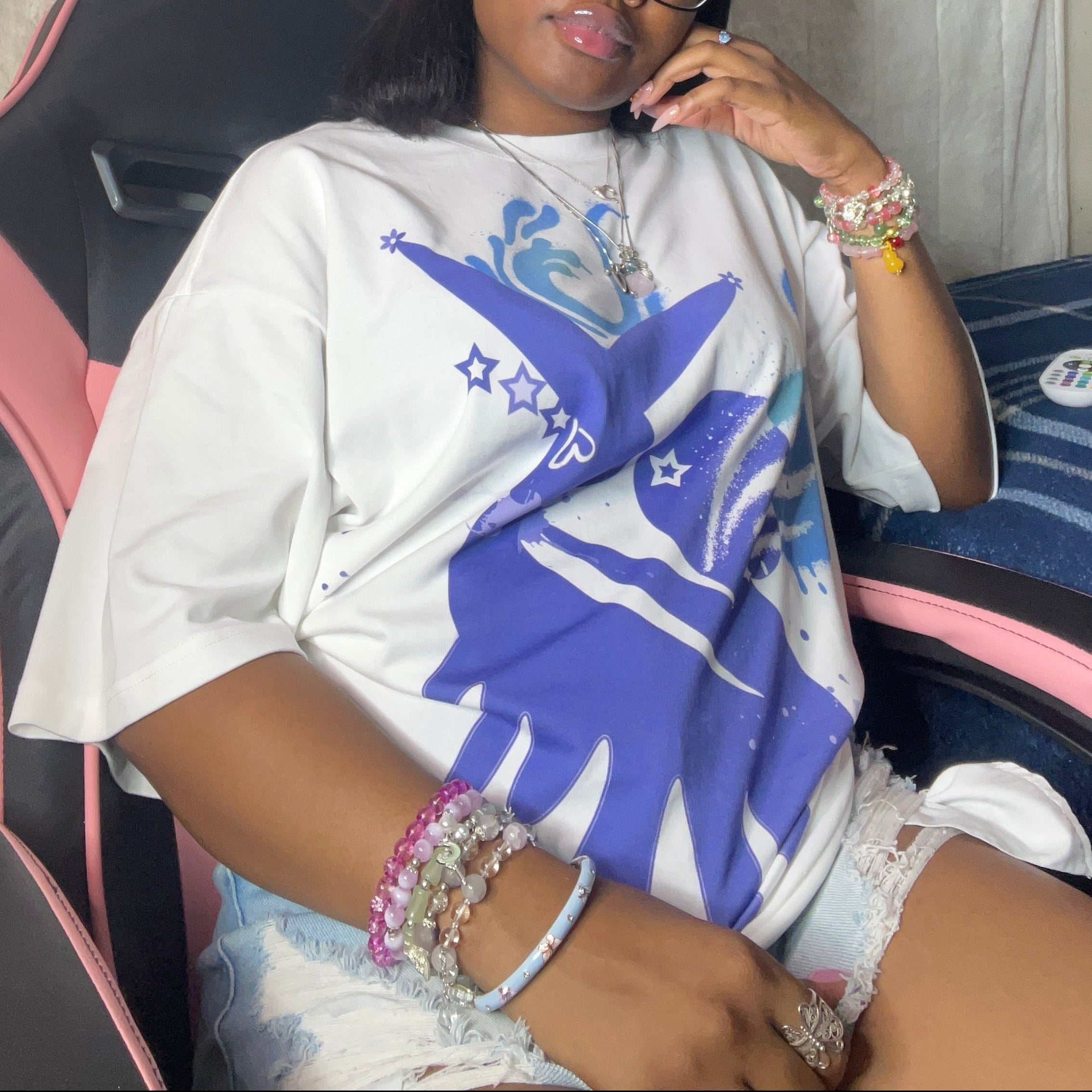 A woman wearing a white graphic t-shirt with a blue and purple design, paired with white shorts, sitting in a pink and black gaming chair against a wall decorated with posters and a yellow pom-pom.