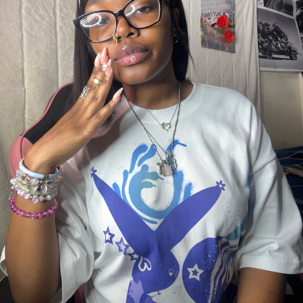 A woman wearing a white graphic t-shirt with a blue bunny design, black-rimmed glasses, and multiple necklaces and bracelets, posing in front of a wall with posters.