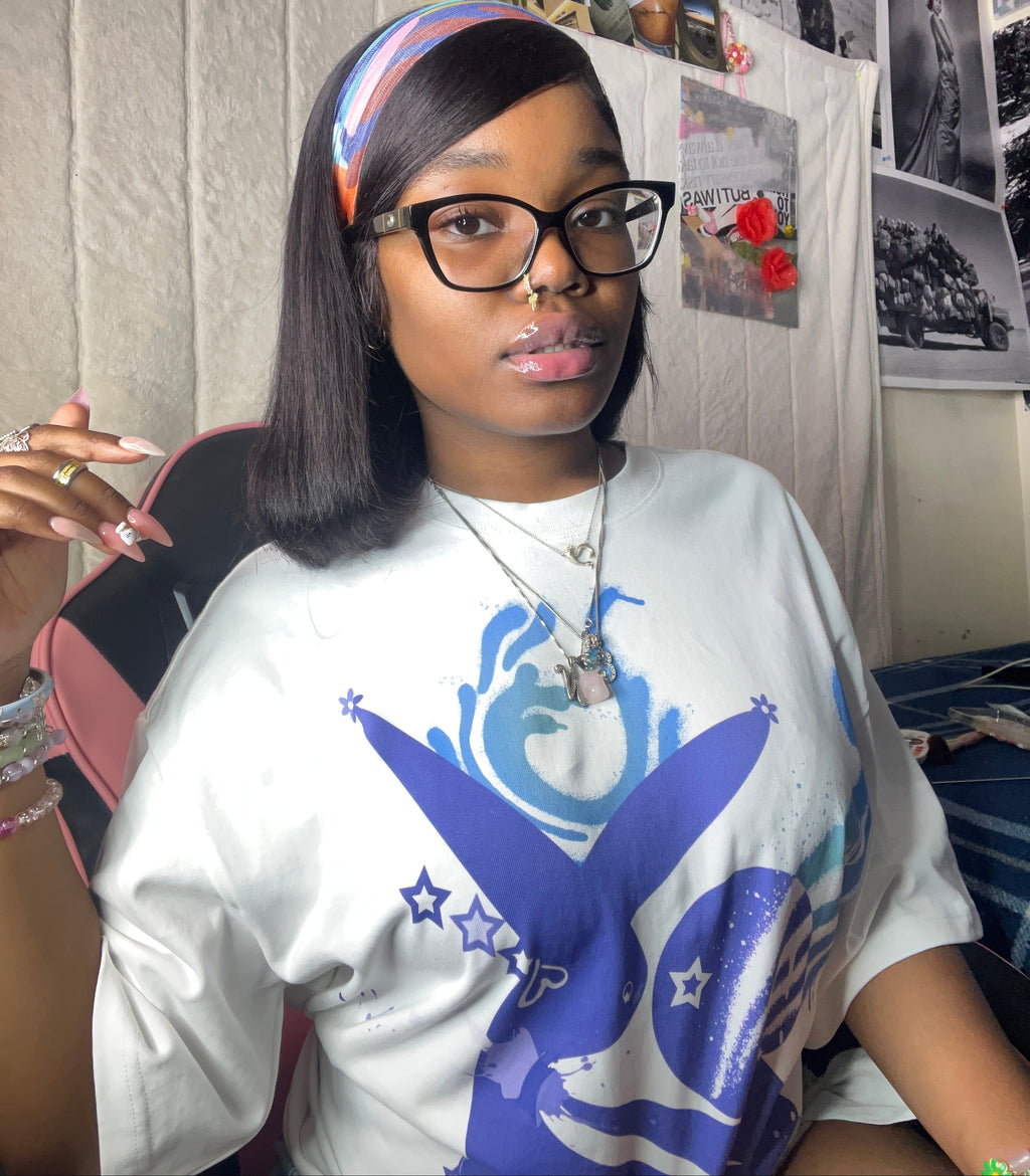 A woman wearing a white graphic t-shirt with a blue and purple bunny design, black glasses, and a colorful striped headband, sitting in a room with posters on the wall.