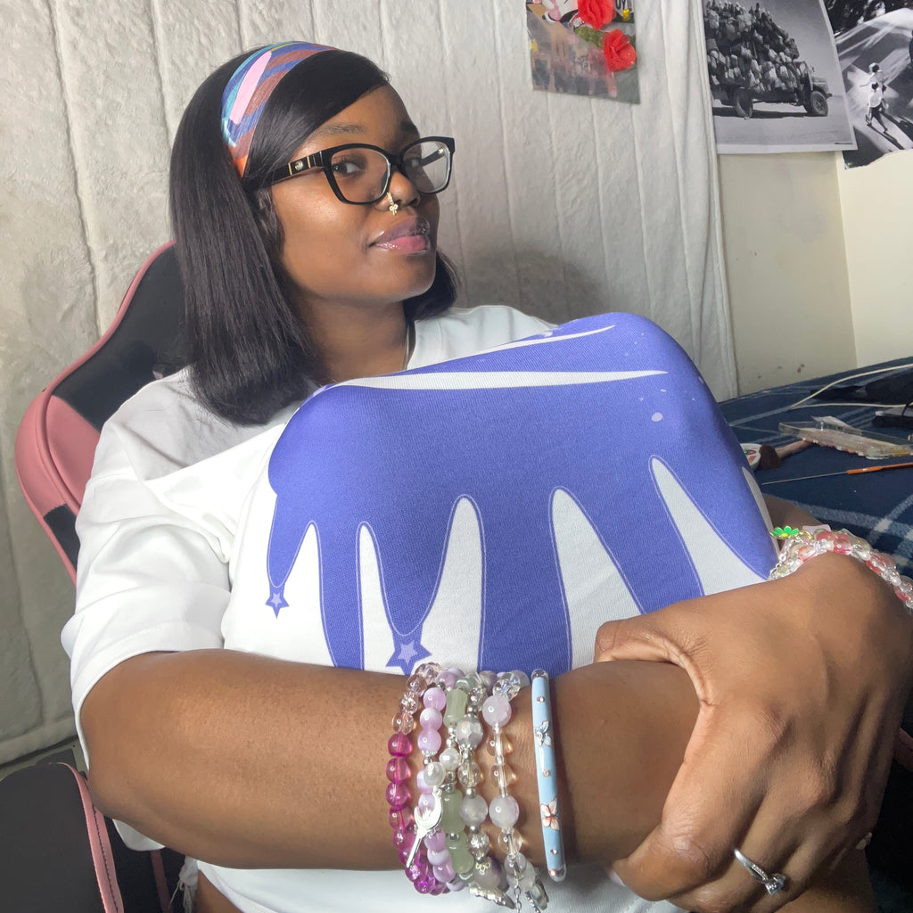A woman wearing a white t-shirt with a blue and white dripping paint graphic sits in a pink and black gaming chair. She is wearing a colorful striped headband, black-rimmed glasses, and multiple beaded bracelets on her wrist.