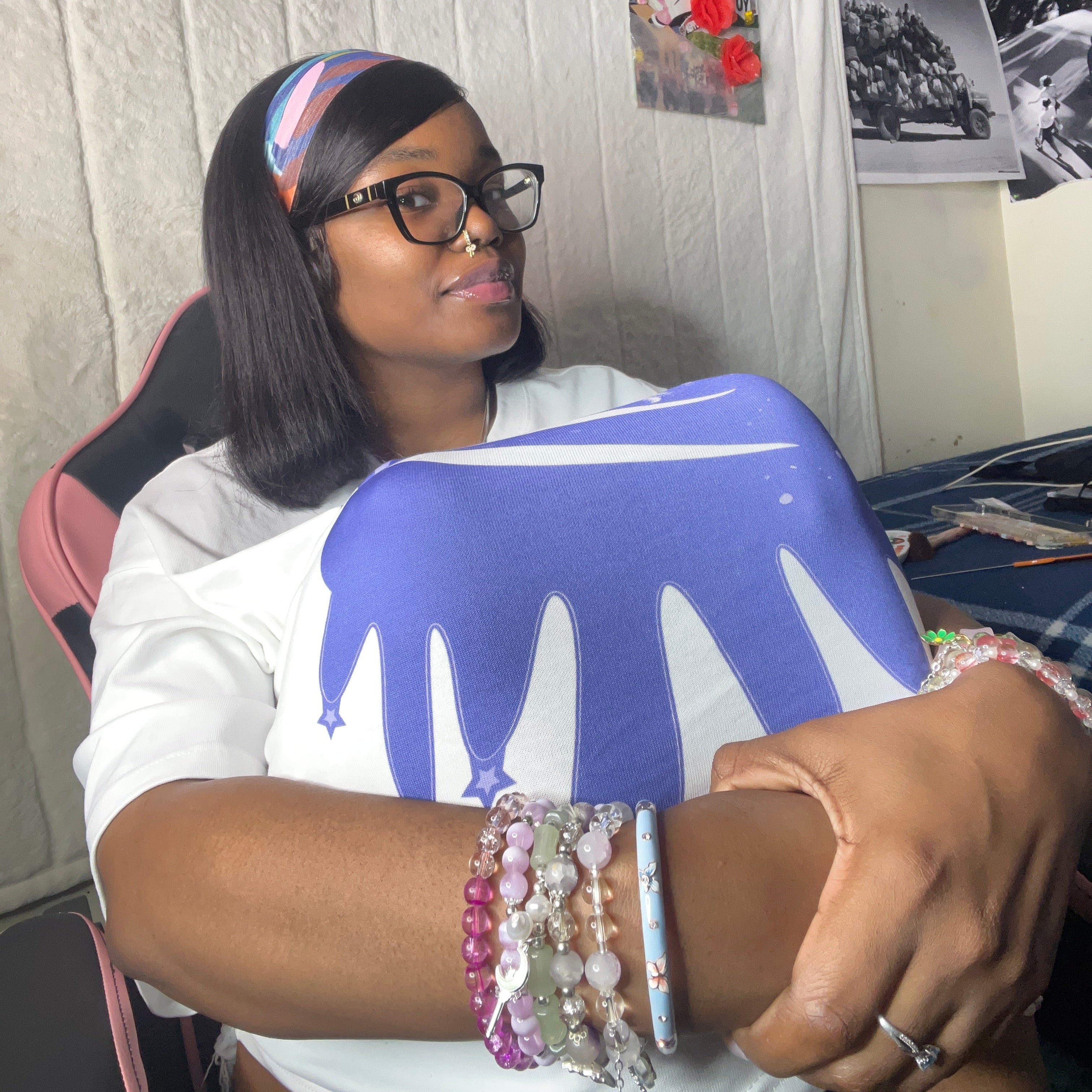 A woman wearing a white t-shirt with a blue and white dripping paint graphic sits in a pink and black gaming chair. She is wearing a colorful striped headband, black-rimmed glasses, and multiple beaded bracelets on her wrist.