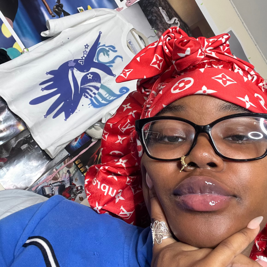 A close-up selfie of a person wearing a red and white Louis Vuitton monogram headwrap and a blue top, with a white Louis Vuitton t-shirt hanging in the background.