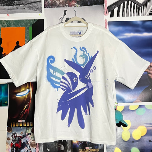 A white short-sleeve graphic T-shirt hanging on a wooden hanger against a collage of posters and photos. The shirt features a large blue and purple abstract bird-like graphic on the front with stars and paint splatter details.